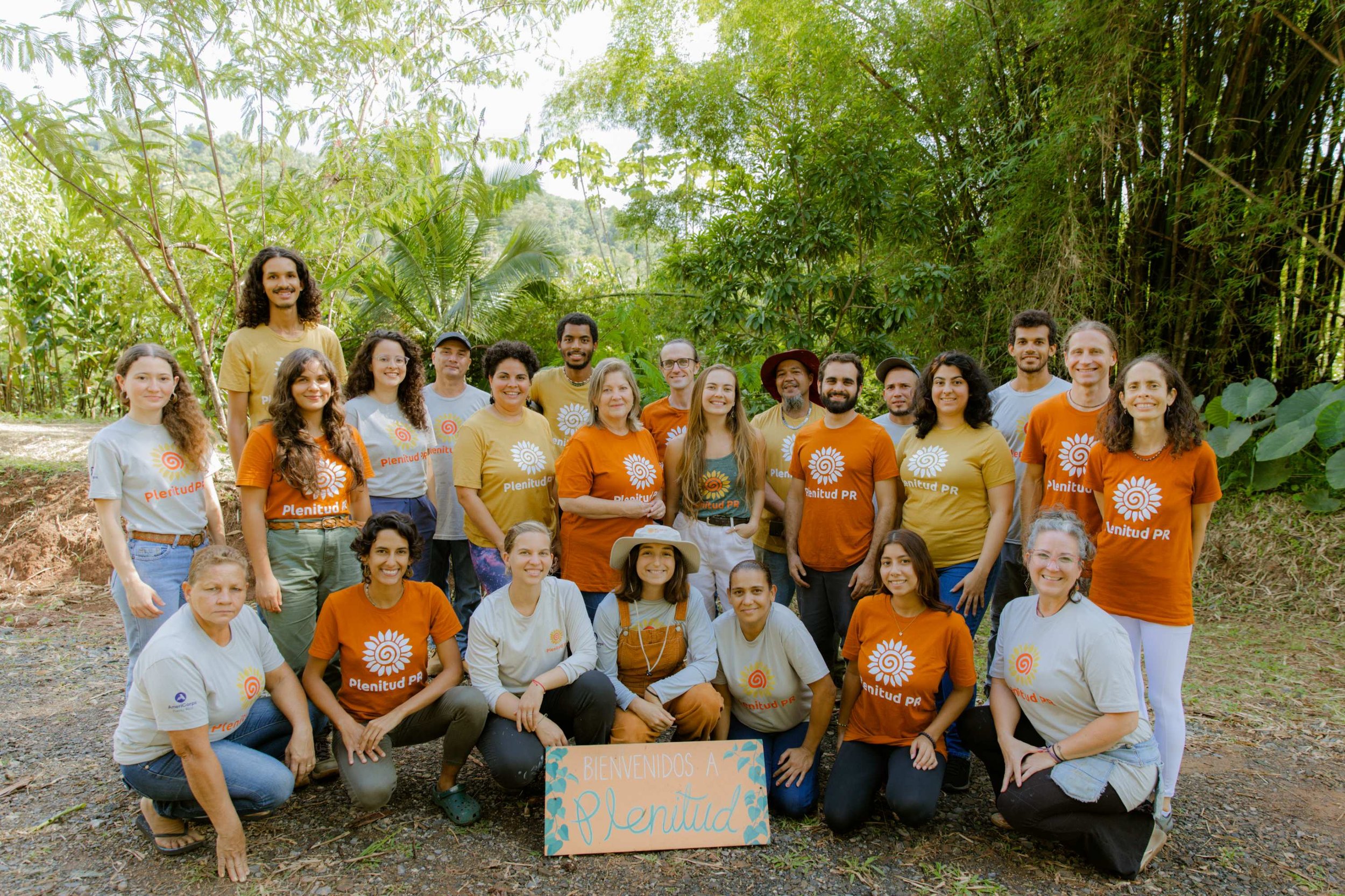 Image of twenty four people standing under light tropical tree cover on woodchip-covered ground. Everyone wears either an orange, yellow, light gray, or light green shirt with Plenitud PR logo, which is a sunflower-like image of swirled circle and petals extending from center. Hand-painted sign at the bottom reads Bienvenidos a Plenitud.