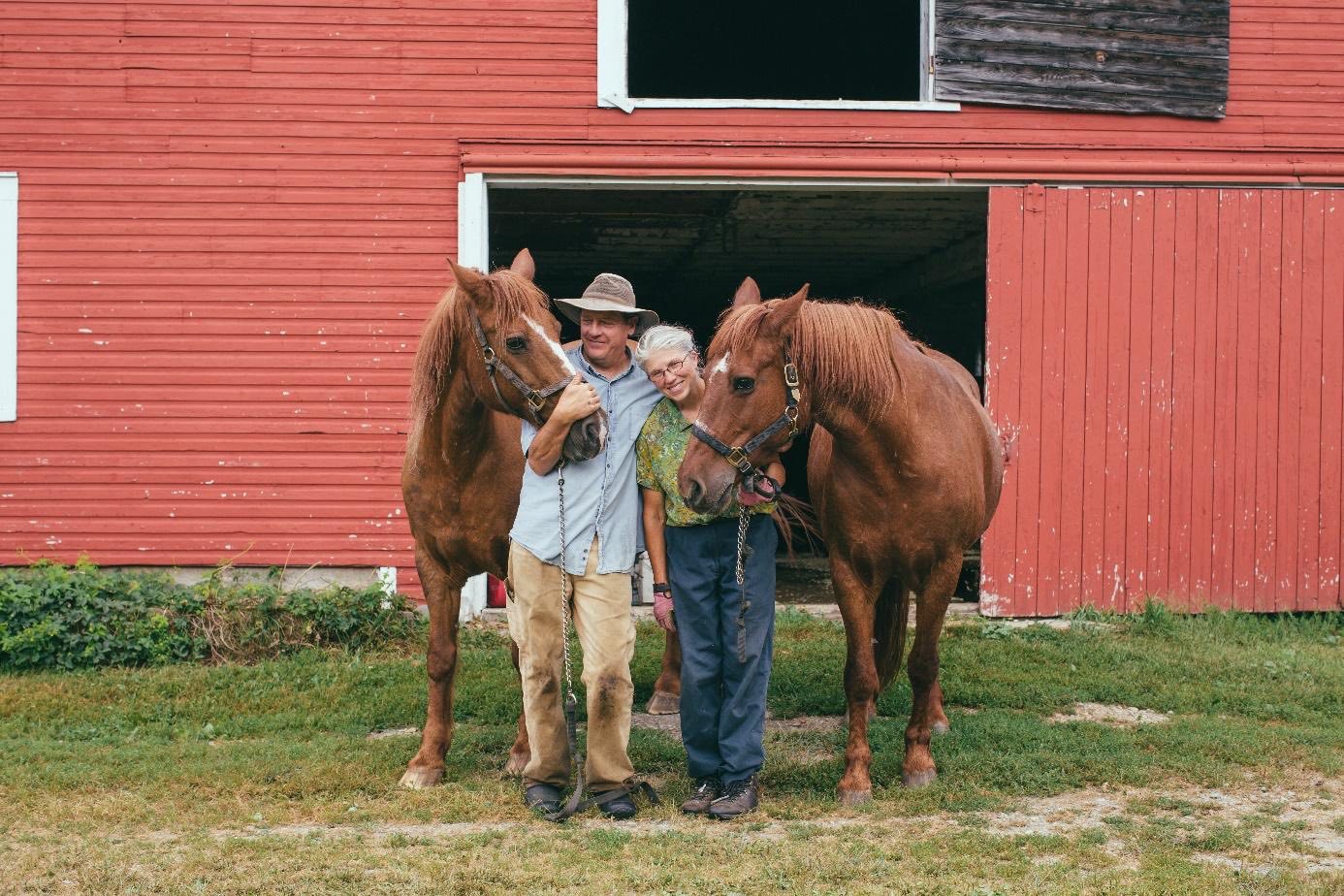 John and Nancy Hayden stand together smiling and wearing work pants and boots hugging two horses standing on either side of them